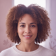 A diverse woman in her late 20s or early 30s, smiling gently, against a blurred backdrop with soft rose and lavender tones.